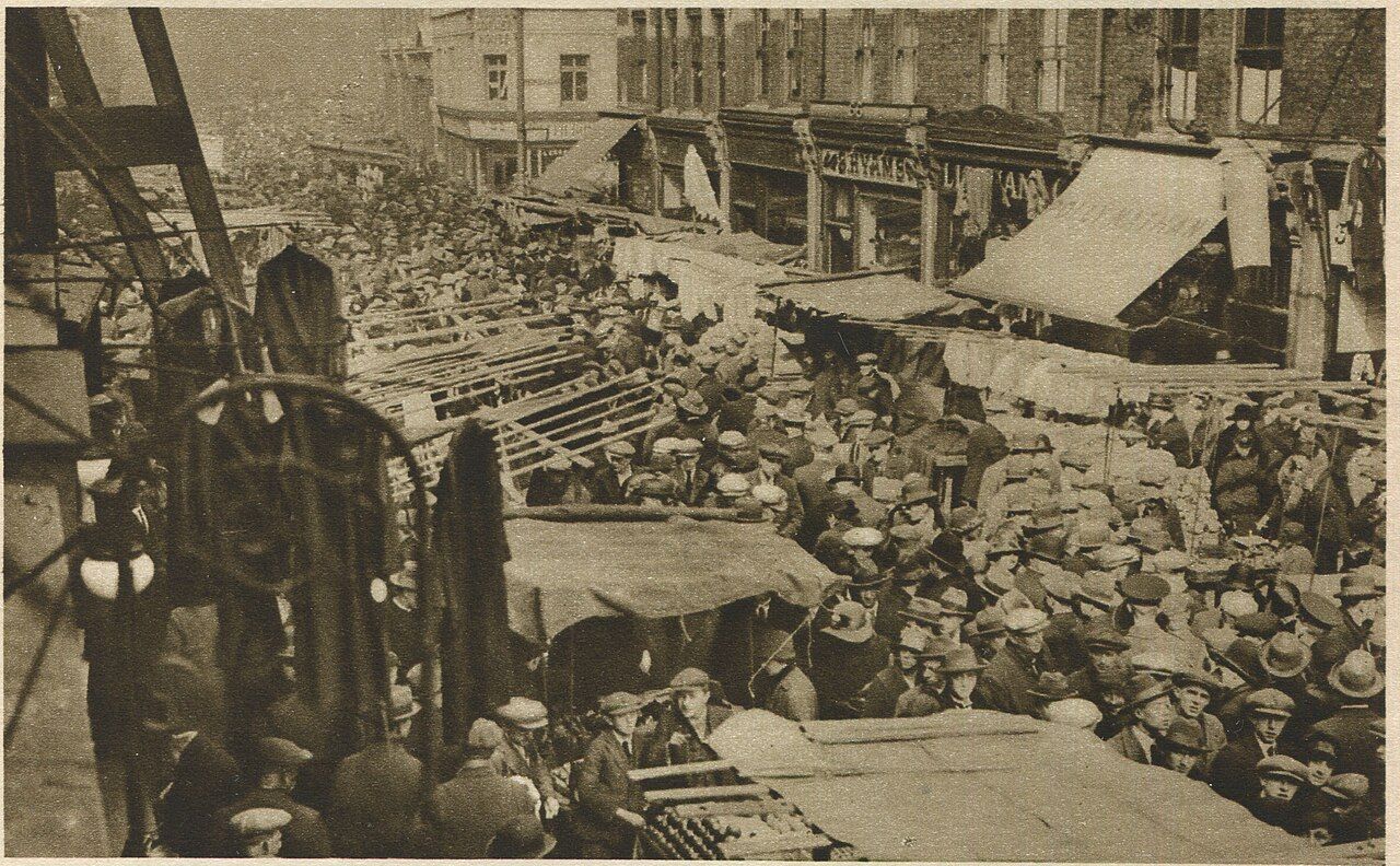 A busy street market on Petticoat Lane, London, 1927, with stalls and crowds.